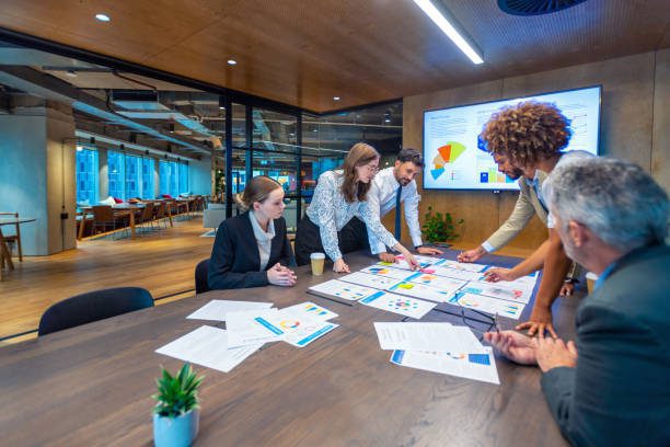 Paperwork and group of peoples hands on a board room table at a business presentation or seminar. The documents have financial or marketing figures, graphs and charts on them. People are pointing at the data. There is a T.V. with financial data in the background